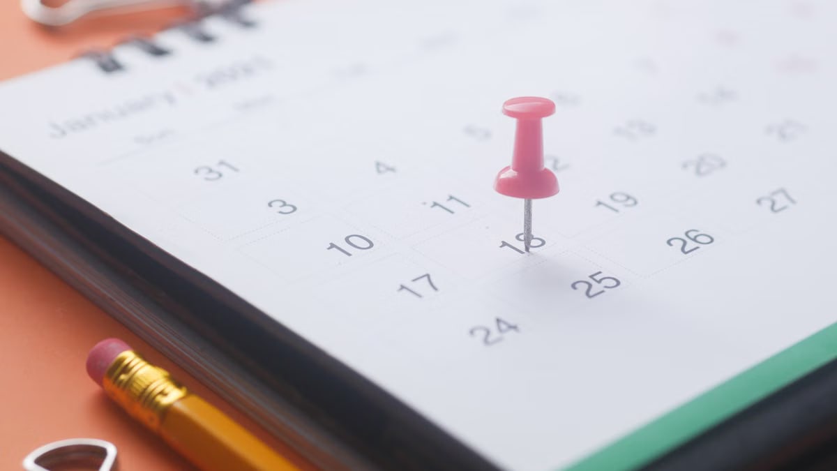 A close-up of a calendar page with a pink push pin, pencil, and binder clip on an orange background.